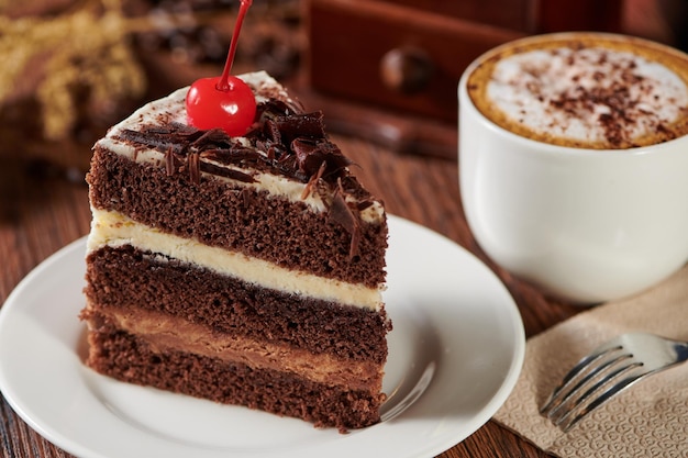 Slice of triple chocolate sponge cake with a cherry on top with a cappuccino on a wooden table