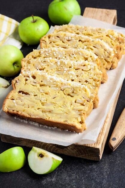 Sliced apple and coconut oaf cake on wooden cutting board on dark background