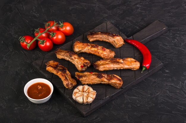 Sliced baked pork ribs on a dark board with hot pepper, garlic and red sauce