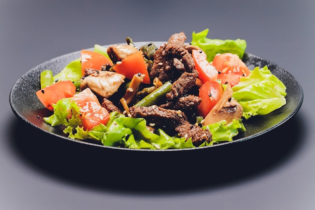 Sliced beef tagliata salad with green beans, cherry tomatoes, fresh arugula and parmesan and lime wedges, served on a black plate.