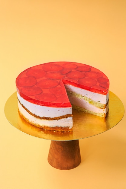 Sliced Birthday Cake decorated with red jelly and strawberries on top on the wooden cake stand. Beautiful sponge cake with whipped cream. Yellow background. Copy space. Food photography for recipe.