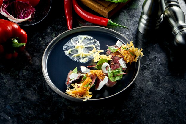 Sliced black roast beef with parmesan chips, coconuts and white sauce, served in a black plate on a dark marble table. Restaurant food