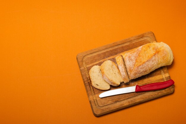 Sliced bread baguette on a wooden cutting board with knife on a terracote color background