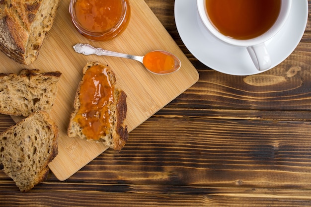 Sliced bread with apricot jam on the cutting board and tea in the white cup on the brown wooden background.Top view.Copy space.