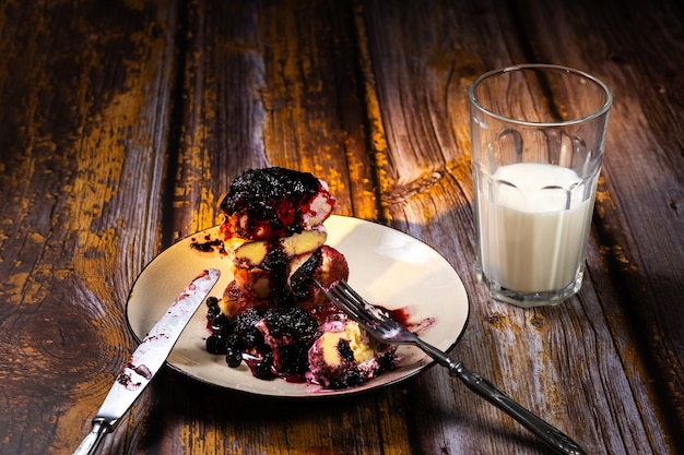 Sliced cheesecakes with blueberry jam and sour cream on a plate and a glass of milk on a wooden table