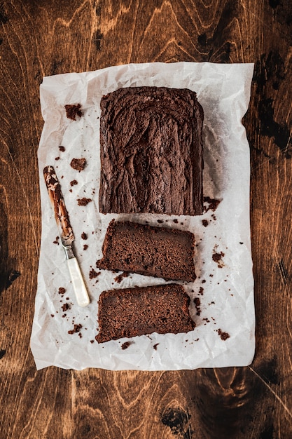 Sliced chocolate loaf cake and a knife on baking paper on dark wooden table, top view