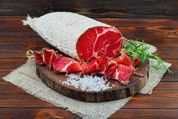 Sliced cured coppa with spices and a sprig of rosemary on dark wooden rustic background.