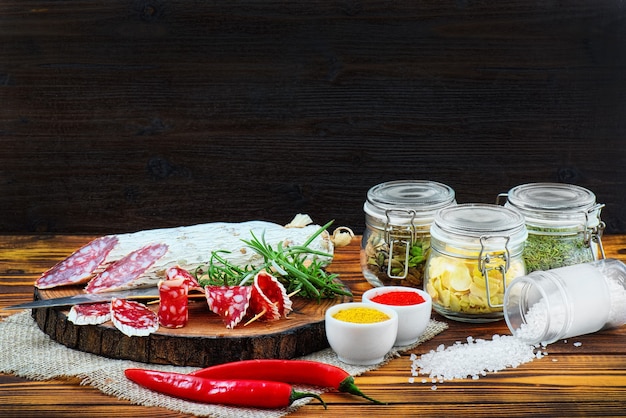 Sliced cured sausage with spices and a sprig of rosemary on dark wooden rustic background.