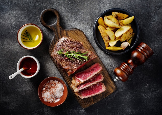 Sliced grilled meat steak New York Striploin with sauce and potato on wooden board on grey background. Top view.