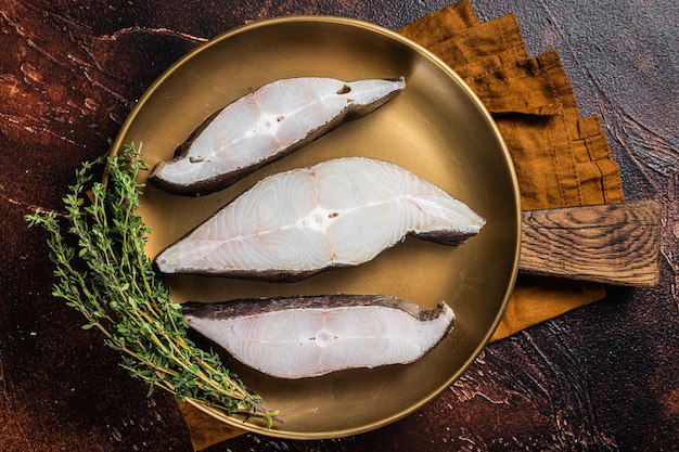 Sliced halibut fish, raw steaks on plate with thyme. Dark background. Top view.