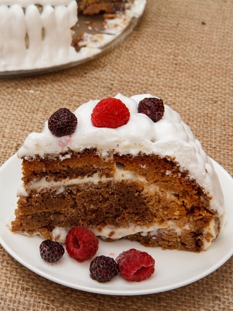 Sliced homemade biscuit cake decorated with whipped cream and raspberries on table with sackcloth.