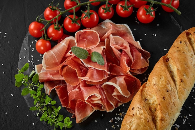 Sliced jamon cherry tomatoes baguette herbs and spices on black stone slate board against a dark grey background Closeup shot Top view