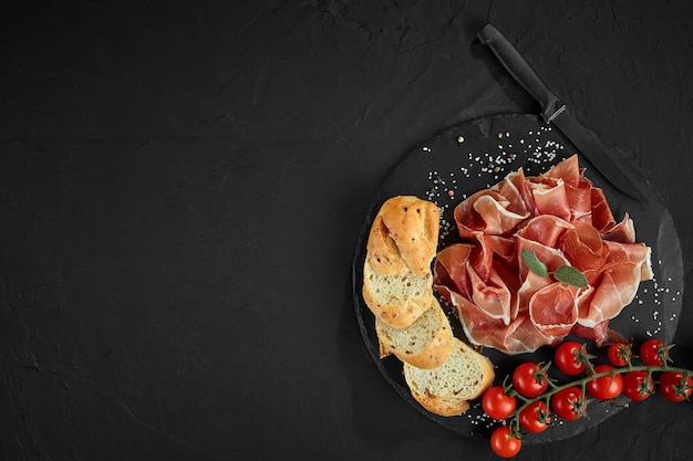 Sliced jamon cherry tomatoes baguette herbs spices and knife on black stone slate board against a dark grey background Closeup shot Top view