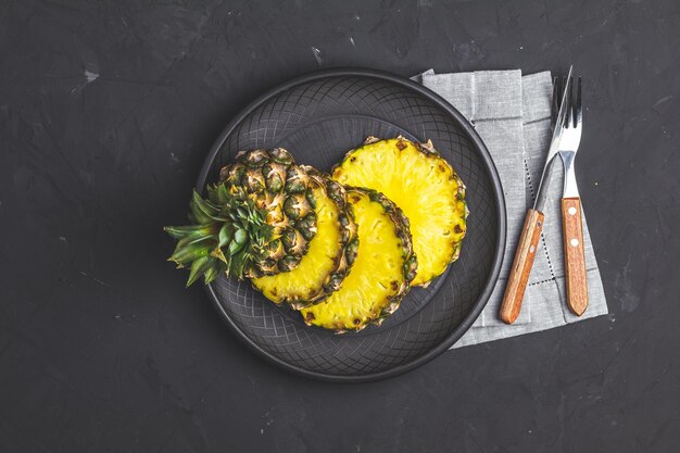 Sliced pineapple in ceramic plate and set of cutlery knife fork on black stone concrete textured surface background Top view with copy space for your text