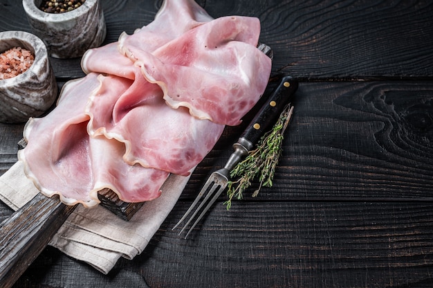 Prosciutto affettato su tavola di legno con erbe aromatiche. fondo in legno nero. vista dall'alto. copia spazio.