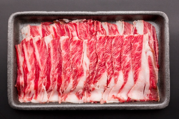 Sliced raw beef place in a row on a black plastic tray isolated on a black background. Sliced ââmeat for cooking, fresh meat for grilling, yakiniku, sukiyaki or shabu.