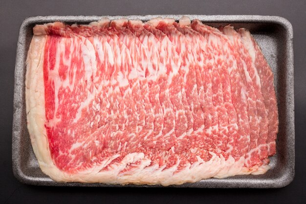 Sliced raw beef place in a row on a black plastic tray isolated\
on a black background. sliced ââmeat for cooking, fresh meat\
for grilling, yakiniku, sukiyaki or shabu.