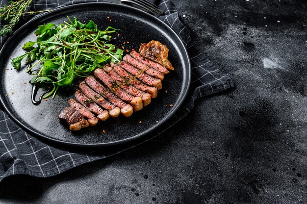 Sliced sirloin steak, marbled beef meat with arugula. Black background. Top view. Copy space