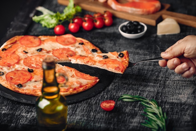 Sliced slice of pizza in hand with cheese, trout, tomatoes, olives and shrimps on chalk board.