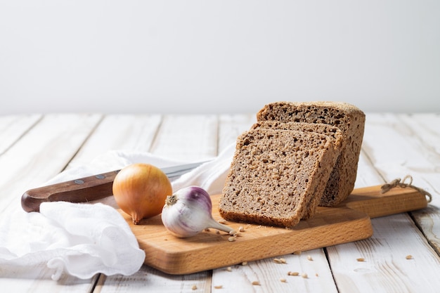 Фото Ломтики хлеба из полбы с луком и чесноком лежат на кухонной доске на светлом деревянном фоне.