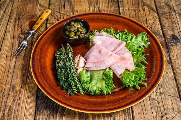 Sliced smoked ham with fresh salad and capers on a plate on wooden table. Top view.