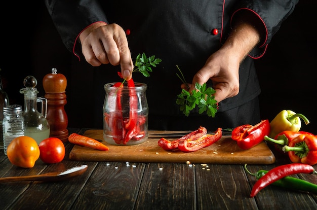 Photo sliced sweet red peppers on a wooden cutting board a professional chef in a black uniform prepares a salad a mans hand puts parsley into a jar