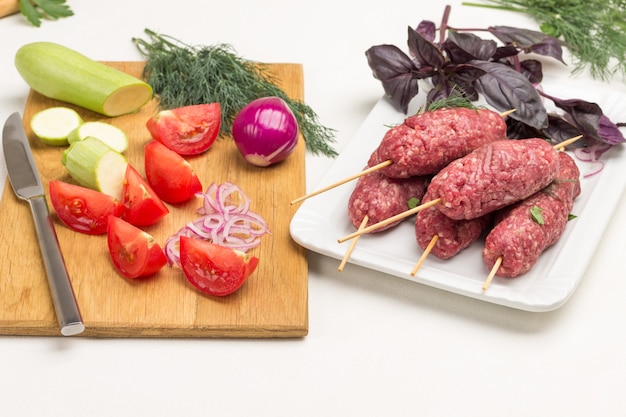 Sliced tomatoes and zucchini on cutting board. Raw minced meat on wooden skewers. Kebab cooking. White background.