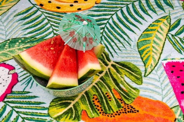 Sliced watermelon with a small red umbrella, on a tropical background of palm leaves.