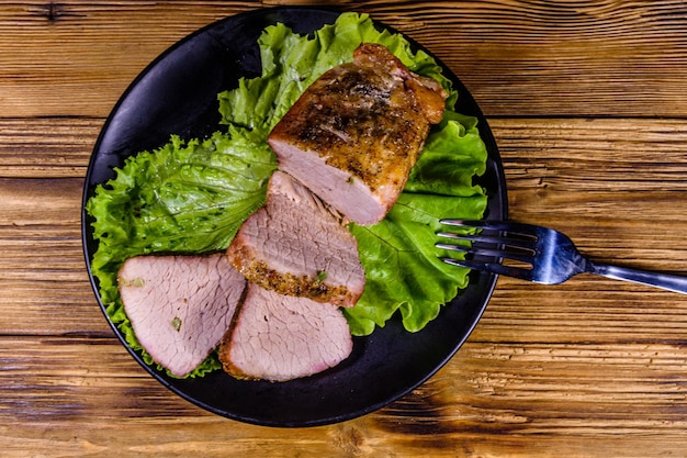 Slices of baked pork meat and lettuce leaves on a black plate Top view