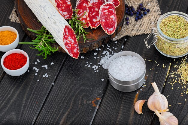 Slices of chopped sausage, spices in jars and scattered, pepper chili and tomatoes on a dark wooden rustic background.