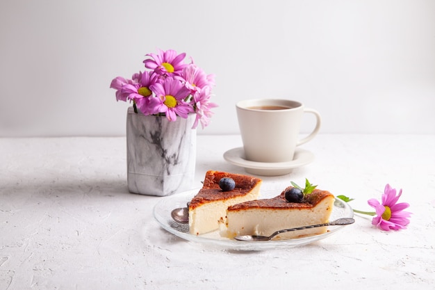 Slices of Homemade Basque burnt cheesecake on plate with blueberries and mint leaves, cup of black tea on light background with flowers in vase