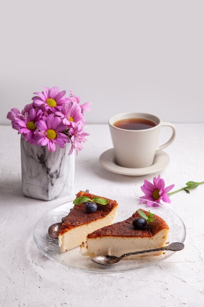 Slices of Homemade Basque burnt cheesecake on plate with blueberries and mint leaves, cup of black tea on light background with flowers in vase