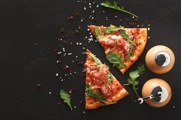 Slices of pizza with prosciutto and rocket salad with spices on
black background, copy space, top view