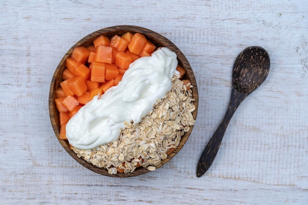 Slices of ripe sweet papaya fruit with oat flakes and white yogurt on coconut bowl on white wooden background close up