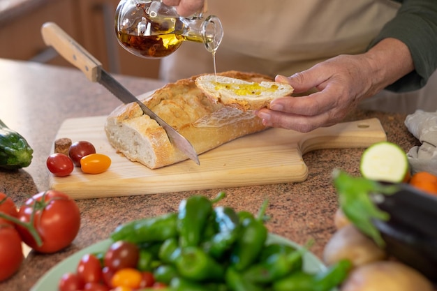 Slices of rustic bread on the wooden cutting board and two woman's hands preparing a snack with a tomato bruschetta and spicy oil. Assortment of fresh vegetables on the table