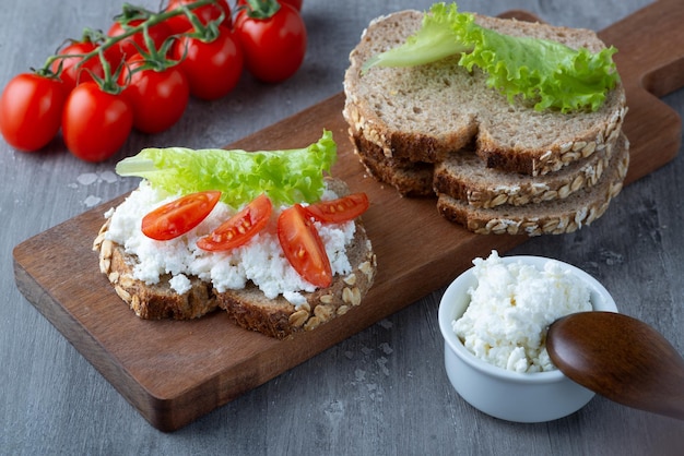 Slices of rye bread with cottage cheese and tomatoes on wooden plate on grey background