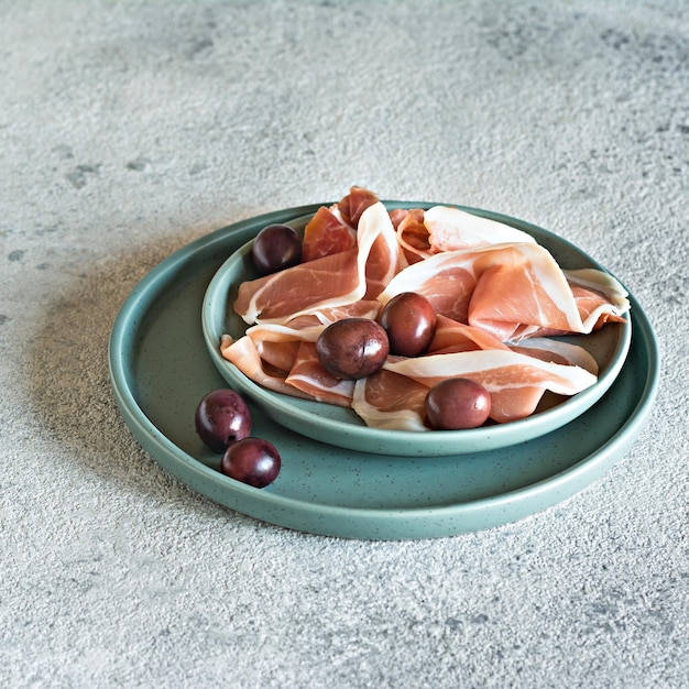 Slices of thinly sliced ham prosciutto Parma ham with a bowl of olives on a light gray background