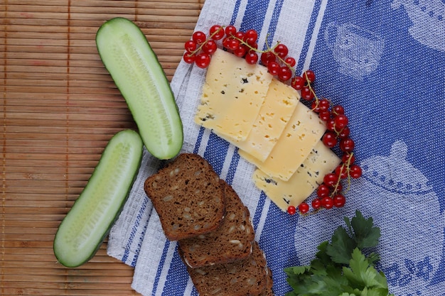 Slices of yellow hard cheese with red currants cucumber and grain bread on a blue napkin Closeup of a healthy snack Healthy food concept