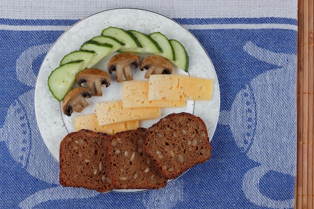 Slices of yellow hard cheese with slices cucumber slices of fried mushrooms and grain bread on white plate on a blue napkin Closeup of a healthy snack Healthy food concept
