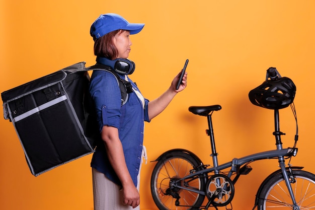 Slide view of takeaway delivery worker standing beside bike\
while checking directions and order details on smartphone.\
restaurant worker wearing delivery uniform while carrying takeout\
food backpack
