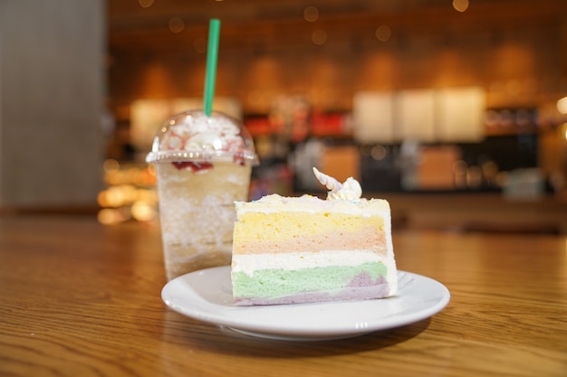 Slided rainbow cake decorate with unicorn horn on top next to coffee frappe. The cake is in a white disk on the wooden table