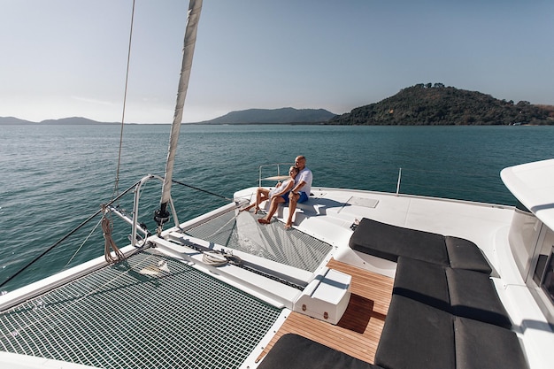 Slightly distant photo of a pleasant couple sitting in an embrace with each other and watching the views passing by, riding on their own white yacht in the middle of the sea. Laxury Concept