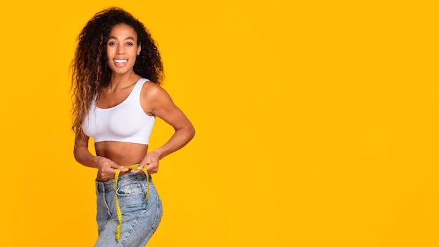 Slim african american woman measuring waist with tape yellow backdrop
