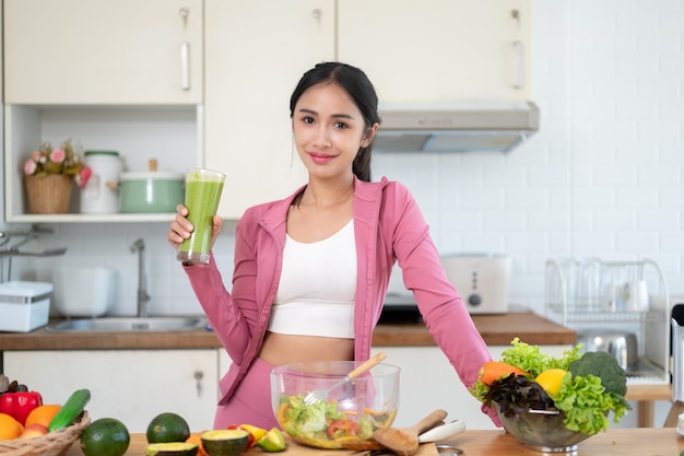 Foto una donna asiatica magra in abiti da palestra tiene in mano un bicchiere di un sano frullato verde al tavolo della cucina