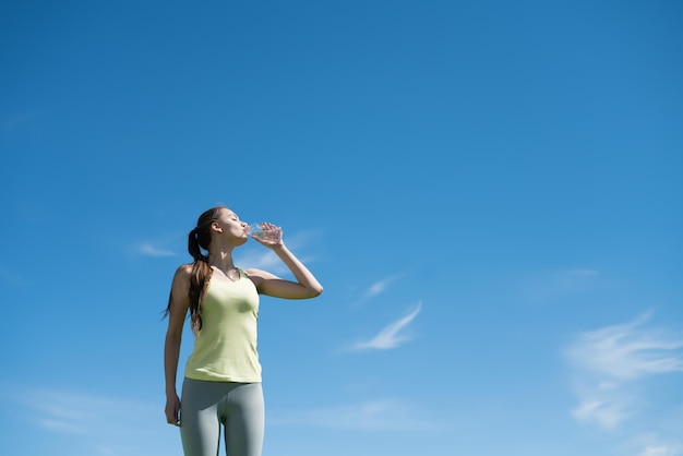 Стройная спортивная девушка пьет воду после тренировки под голубым небом