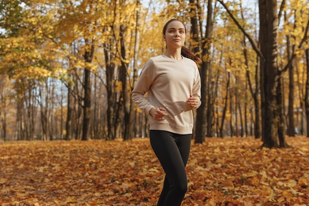 Slim athletic woman in sportswear running in autumn park on sunny day during active training