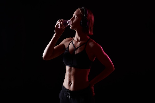 La donna magra e attraente in abiti sportivi con le cuffie beve acqua dopo l'allenamento su sfondo nero isolato