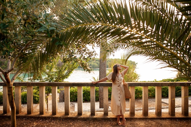 Slim brunette dressed in light sundress and sandals leaning on balusters and looking up under large palm leaf Fit lady with straw bag in hand exploring Brac Island during summer vacation in Croatia