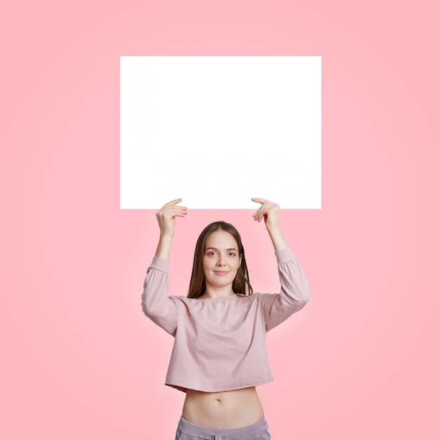 Photo slim caucasian woman in casual clothes, holds white blank white card for your advertisement or promotional content, isolated on pink. free space. vertical shot.