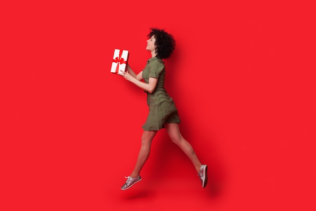 Slim caucasian woman with curly hair running on a red studio wall with a present giving it to somebody